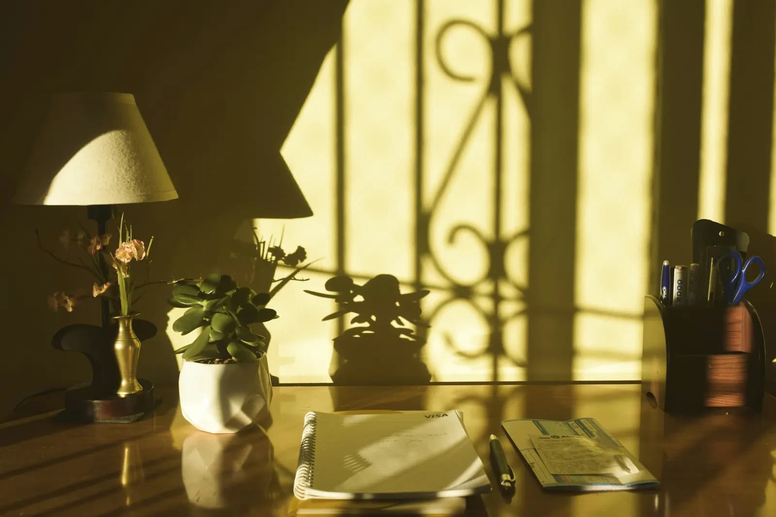 A wooden desk in late afternoon light with a table lamp, a small plant, a notebook and a pen