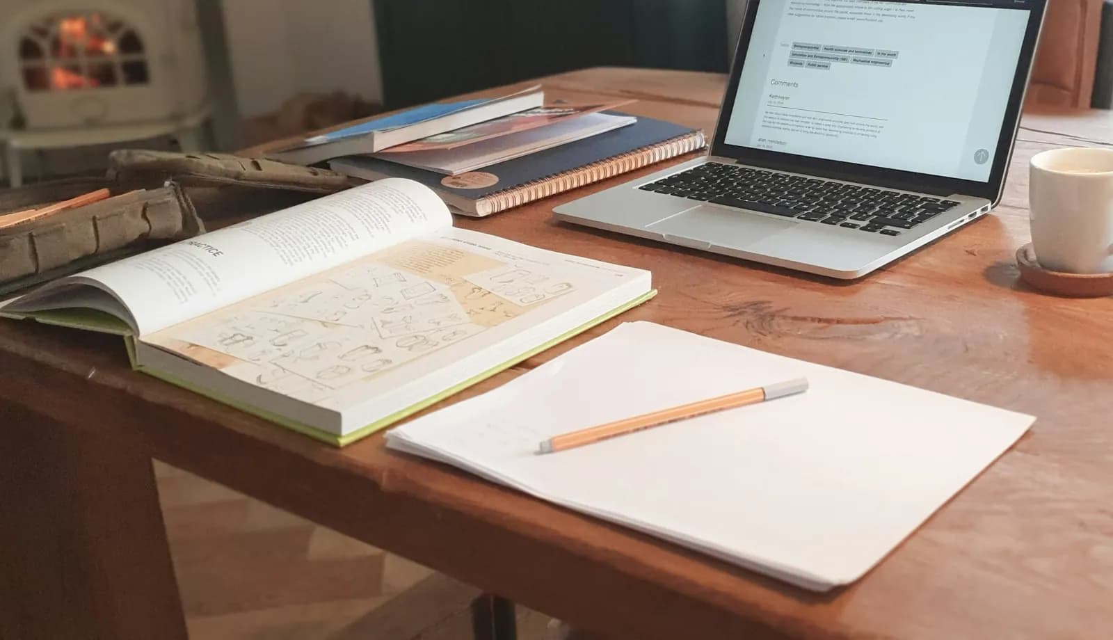 A laptop open on a wooden table at home beside an open book, a notepad and a pencil