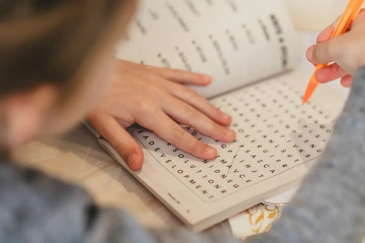 A child's hands resting on a word puzzle page while holding an orange pencil