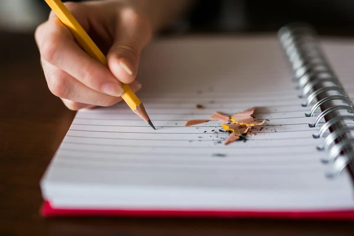 A hand holding a yellow pencil over a spiral notebook, with pencil shavings scattered on the page