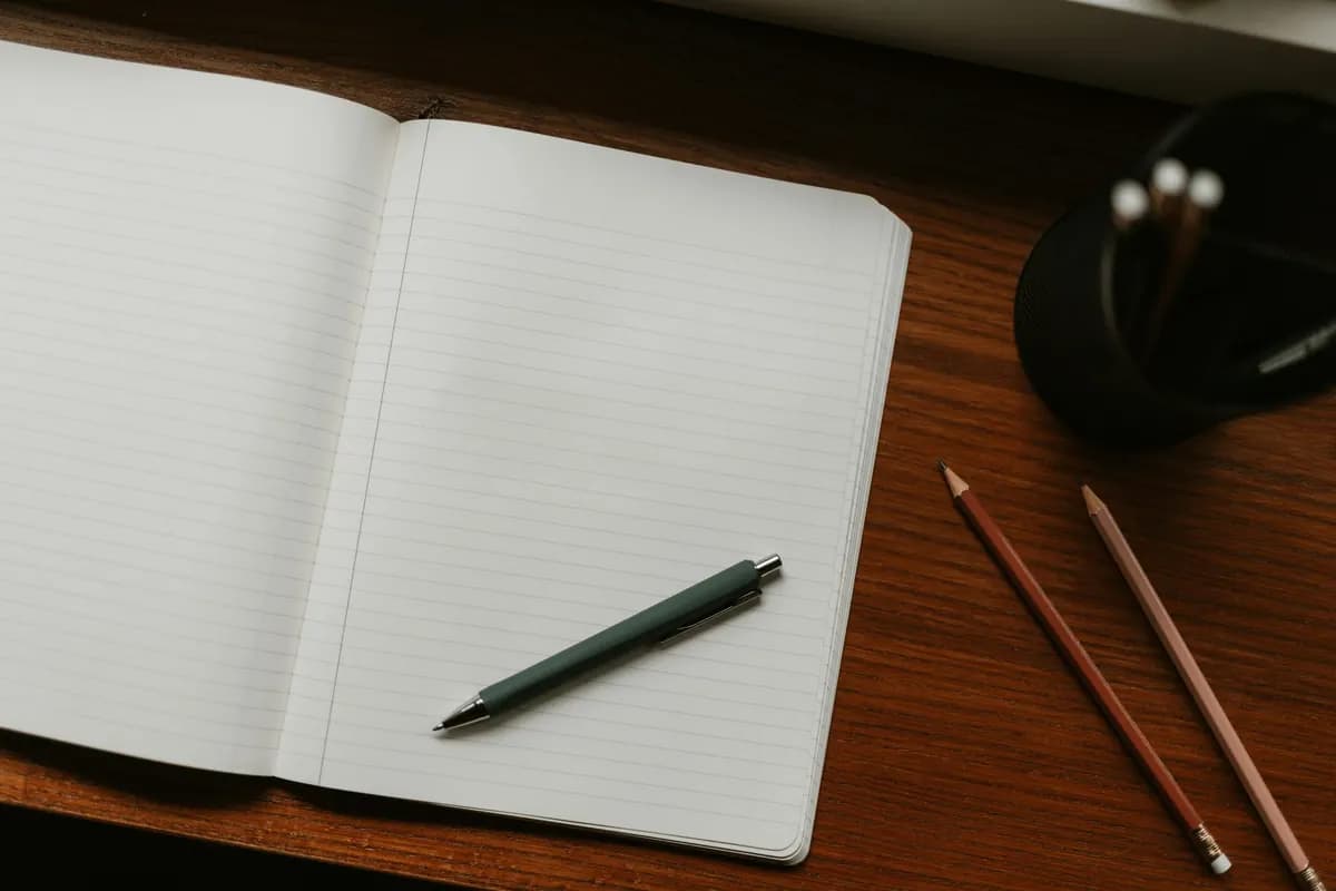 An open lined notebook on a dark wooden desk with a pen across the page and two pencils beside it