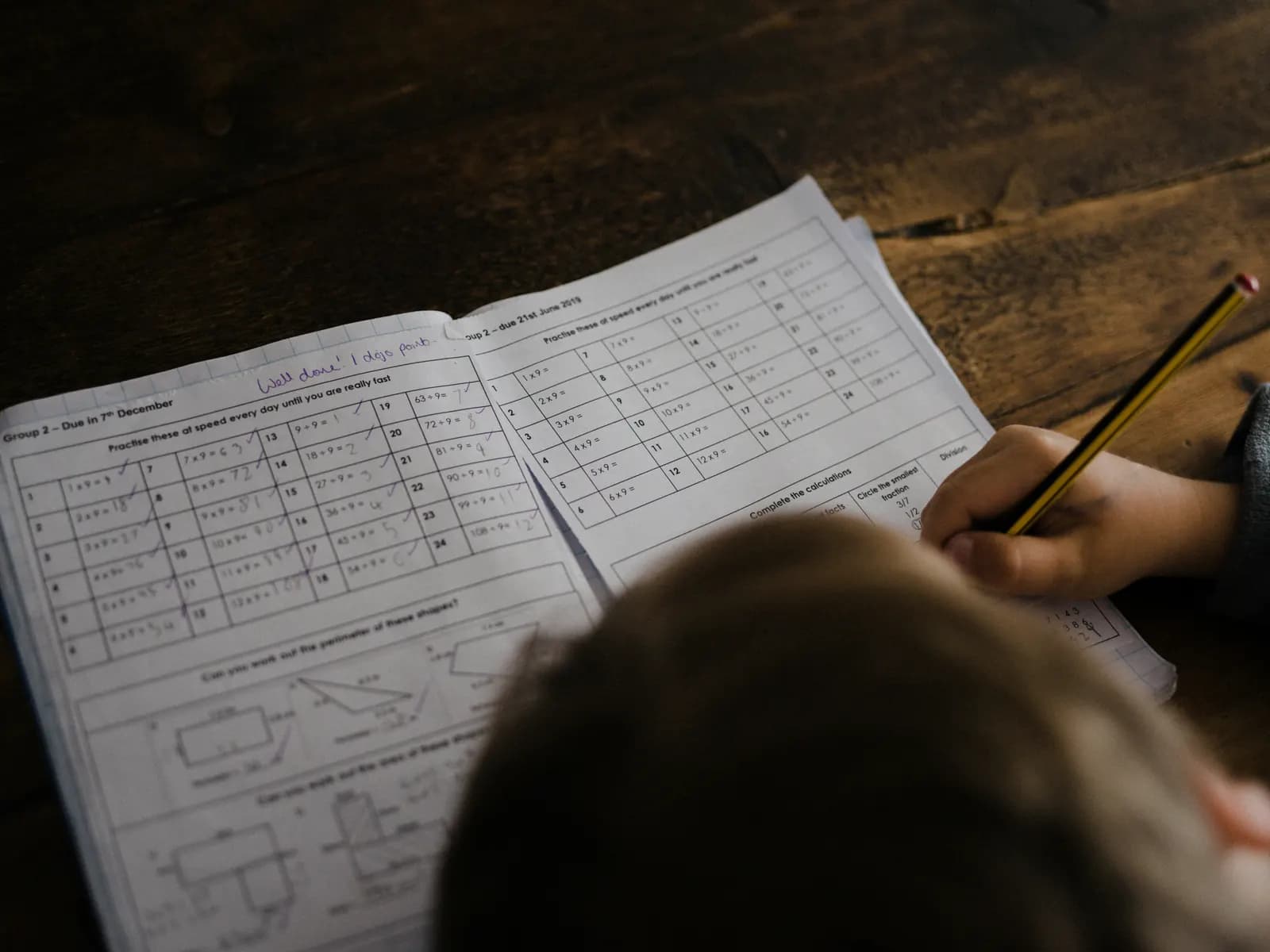 A child working through a times tables homework sheet with a pencil, seen from behind